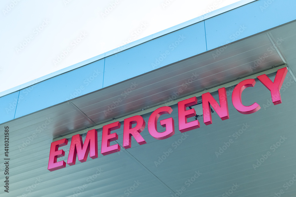 An emergency sign outside a hospital. The illuminated red letters spell emergency and are affixed to a grey metal panel overhang. The signage is in capital letters. It's a medical hospital department. An emergency sign outside a hospital. The illuminated red letters spell emergency and are affixed to a grey metal panel overhang. The signage is in capital letters. It's a medical hospital department.
