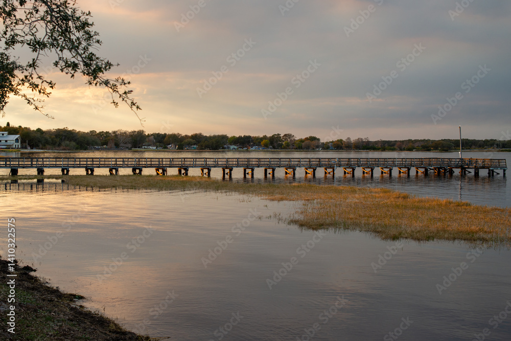 Obraz premium A pier, raised over a calm body of water and juts out from the shore as the evening sun goes down. The wooden structure allows people to fish, moor boats, and dock from the wharf. The sky is golden.