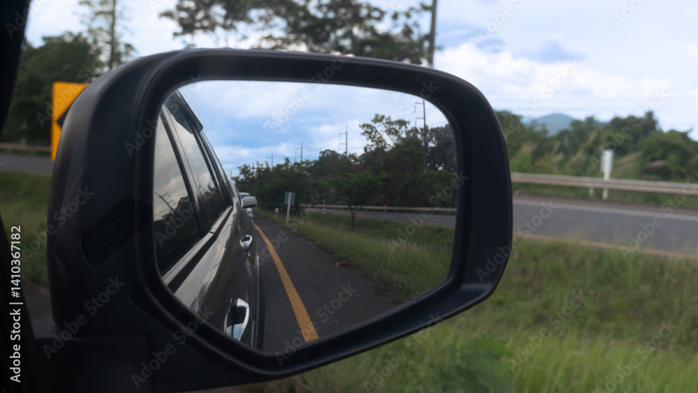 Side view of mirror wing of car grey color. Driving on asphalt road. Side with green grass. Inside mirror wing can see road and trees under blue sky. Side view of mirror wing of car grey color. Driving on asphalt road. Side with green grass. Inside mirror wing can see road and trees under blue sky.