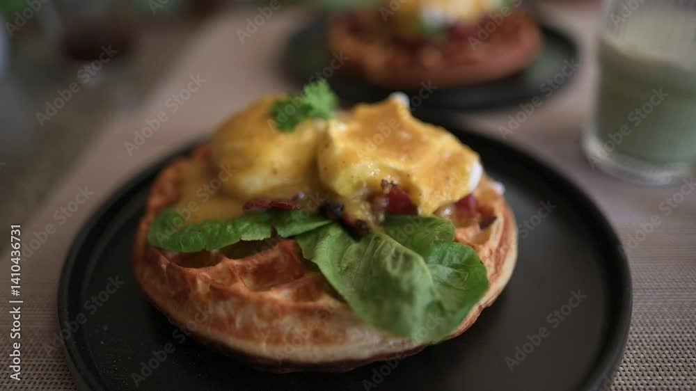 Close-up of eggs benedict on crispy waffle base garnished with fresh greens and hollandaise sauce, served on black plate with blurred matcha drink in background on breakfast table eggs benedict Close-up of eggs benedict on crispy waffle base garnished with fresh greens and hollandaise sauce, served on black plate with blurred matcha drink in background on breakfast table eggs benedict