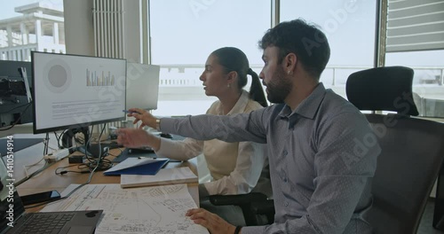 Business colleagues analyze data on computer screens, discussing and pointing at charts, in a modern office setting.