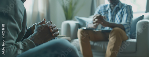 A doctor is taking notes while talking to their patient, who sits on the couch with their hands clasped together and appears distressed. The background shows an office setting with soft lighting.