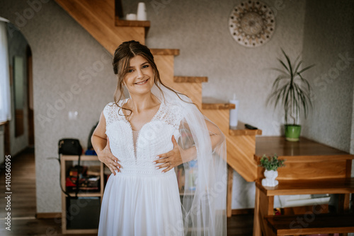 Smiling bride in a white wedding dress adjusting her gown indoors before the ceremony, showing joy and excitement on her special day.. photo