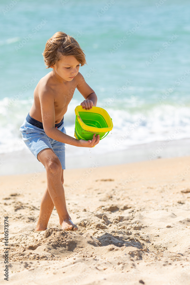 Happy child boy playing with the sand on the beach, pouring water from plastic bucket on the sea beach, building sand castle, enjoying summer. Advertising for tour operators, hotels, airlines.