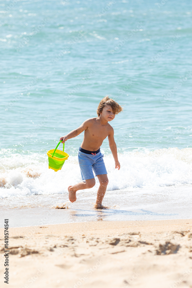 Happy child boy playing with the sand on the beach, pouring water from plastic bucket on the sea beach, building sand castle, enjoying summer. Advertising for tour operators, hotels, airlines.