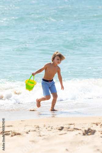 Obraz na płótnie Happy child boy playing with the sand on the beach, pouring water from plastic bucket on the sea beach, building sand castle, enjoying summer. Advertising for tour operators, hotels, airlines. w salonie