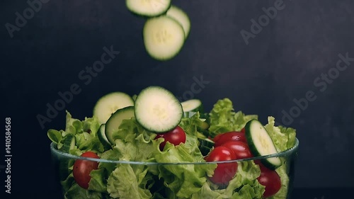 Fresh cucumber slices falling on lettuce and tomatoes on black background. Eating healthy raw organic food, vitamins, vegetables concept. Slow motion