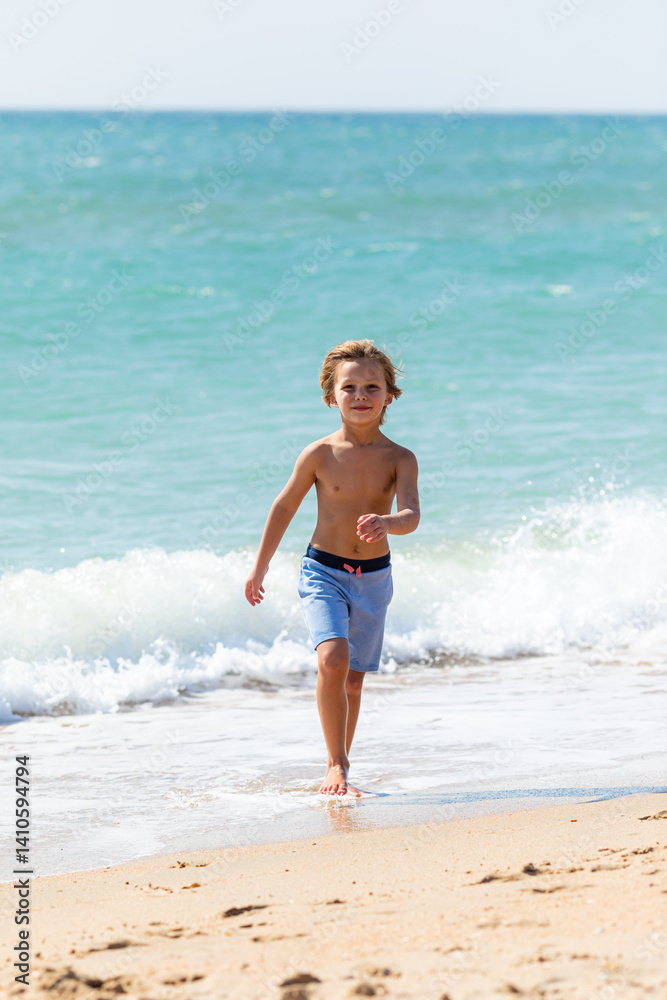 Cute little boy playing on the beach on summer holidays. Joyful child in nature with beautiful sea, sand and blue sky. Happy kid on vacations at seaside running in the water and having fun