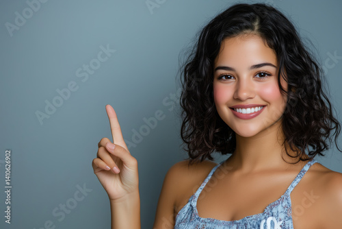 Smiling woman with curly hair posing and pointing up, wearing a patterned top, expressing happiness and positivity in a studio setting with a neutral gray background.