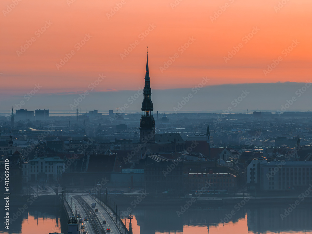 Serene sunrise over Riga, Latvia, featuring St. Peter's Church spire, a bridge over the Daugava River, and reflections in calm water with morning mist.