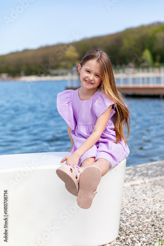 Happy little girl with long blonde hair, in lilac summer dress, sitting on the beach with blue water background. Children's Day. Vacation concept. Space for text. Advertising for tour operators