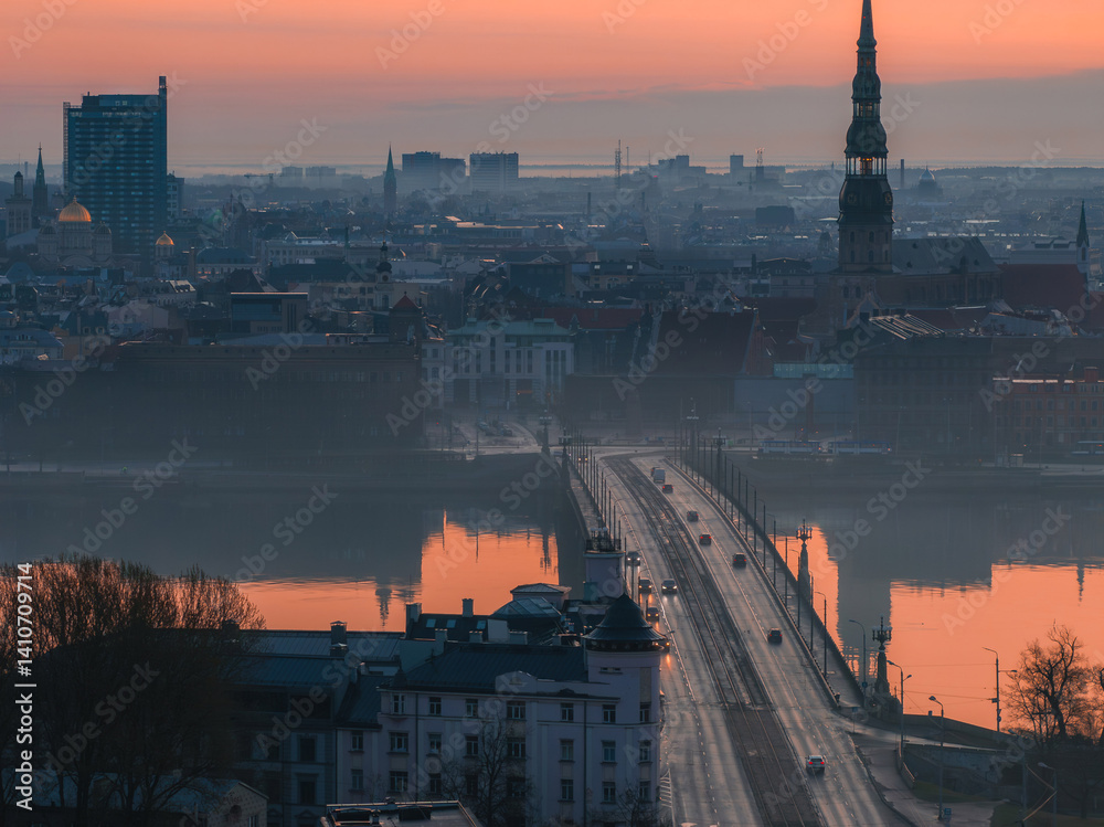 Serene sunrise over Riga, Latvia, featuring St. Peter's Church spire, the Daugava River with a bridge, and a mix of historic and modern buildings.