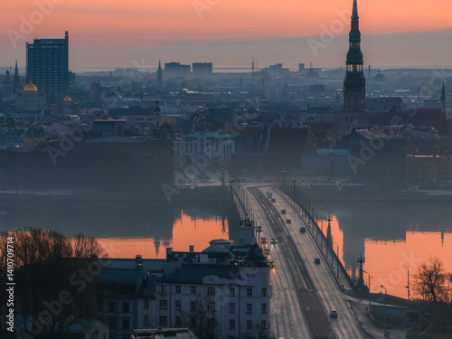 Obraz na płótnie Serene sunrise over Riga, Latvia, featuring St. Peter's Church spire, the Daugava River with a bridge, and a mix of historic and modern buildings. w salonie