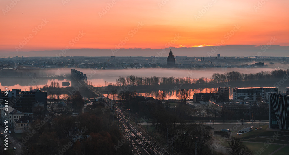 Serene sunrise over Riga, Latvia, featuring St. Peter's Church spire, mist covered Daugava River, a railway bridge, and urban structures in soft light.