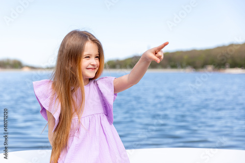 Happy little girl with long blonde hair, in lilac summer dress, sitting on the beach with blue water background and points index finger far away. Vacation concept. Advertising for tour operators