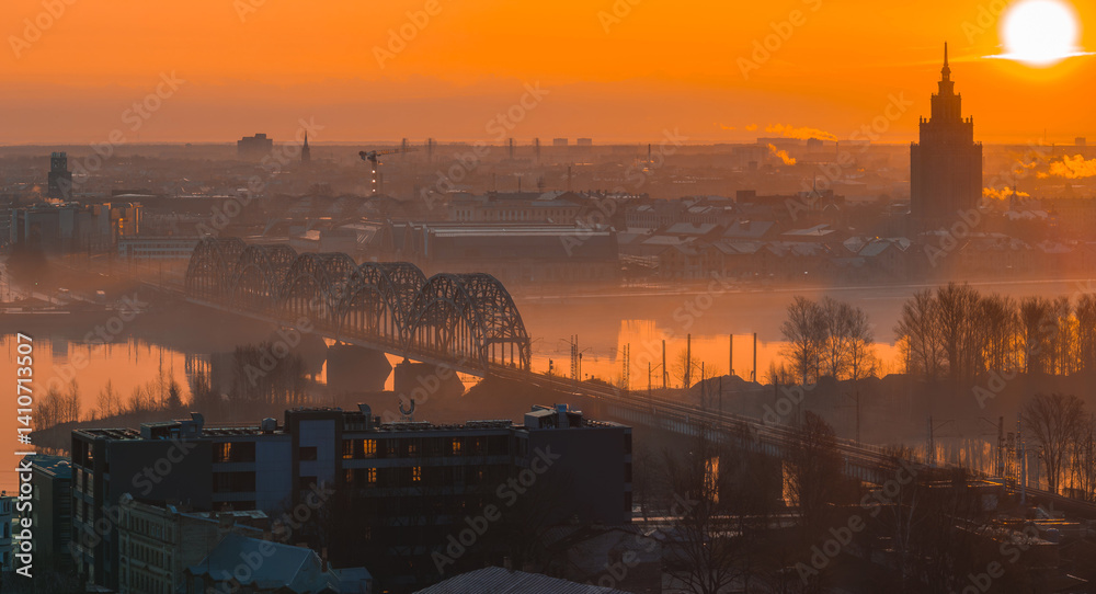 A warm sunrise illuminates Riga, Latvia, with the arched railway bridge over the Daugava River and the Latvian Academy of Sciences in the background.