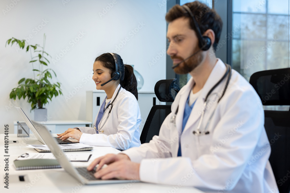Two healthcare professionals wearing headsets and lab coats, working on laptops in a modern office setting. They are smiling while using technology.