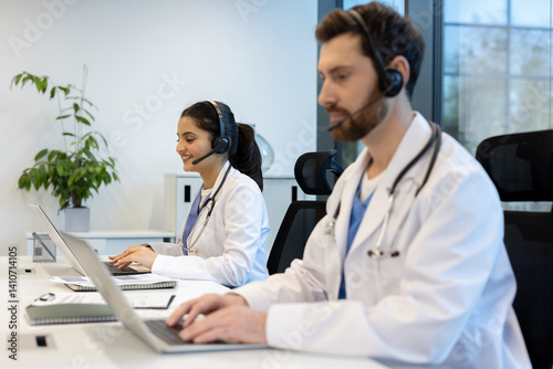 Obraz na płótnie Two healthcare professionals wearing headsets and lab coats, working on laptops in a modern office setting. They are smiling while using technology. w salonie