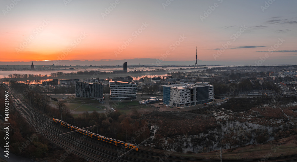 Serene sunrise in Riga, Latvia, featuring the Riga TV Tower, Daugava River, modern buildings, a yellow train, and historic spires in the distance.