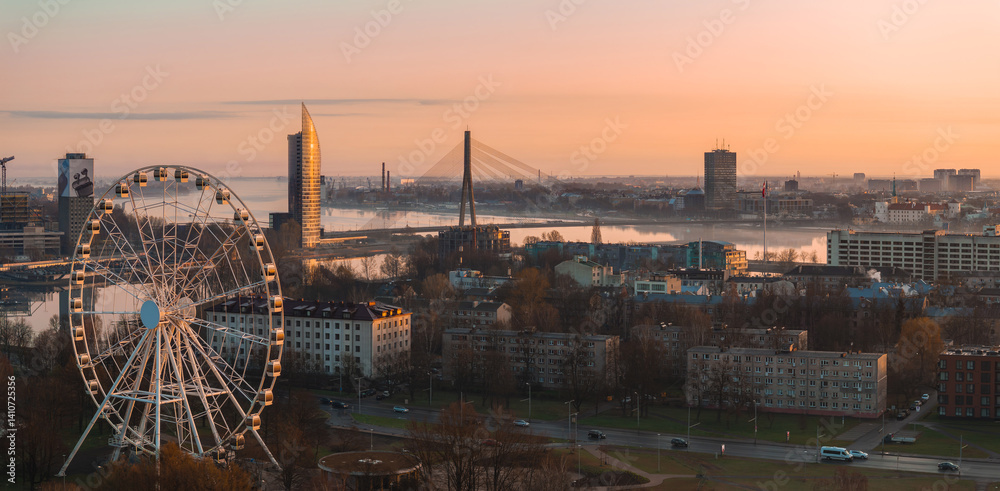 Serene sunrise view of Riga, Latvia, featuring the Vansu Bridge, Z Towers, a large Ferris wheel, and the Daugava River reflecting warm light.