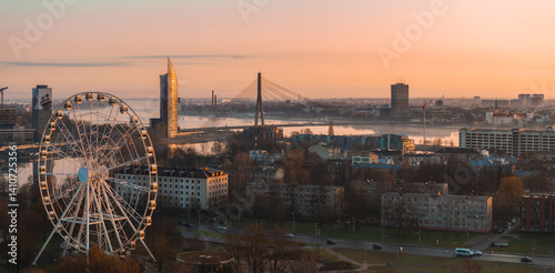 Obraz na płótnie Serene sunrise view of Riga, Latvia, featuring the Vansu Bridge, Z Towers, a large Ferris wheel, and the Daugava River reflecting warm light. w salonie