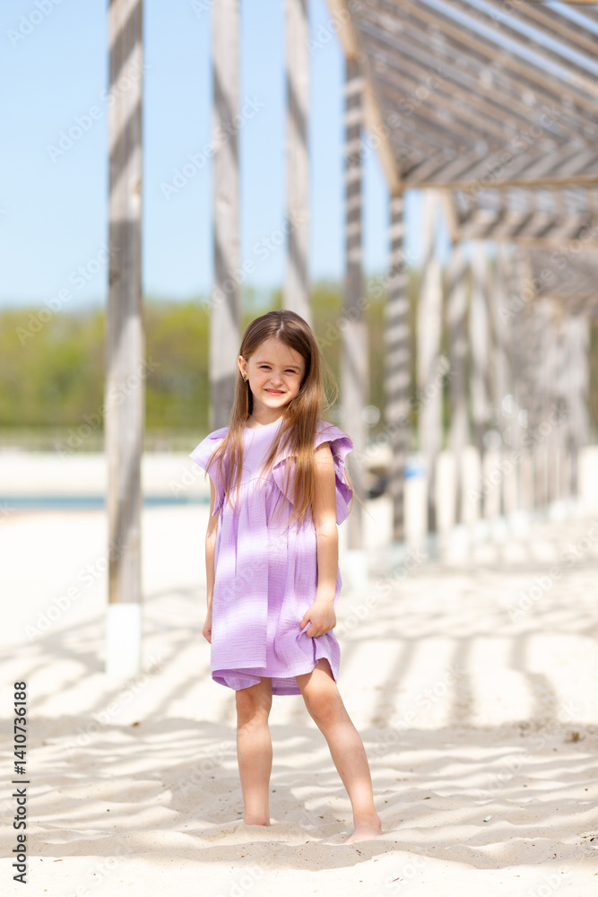 Carefree summer. Little girl in lilac dress has fun and posing on sandy beach under wooden gazebo on sunny day. Joyful childhood adventures and tropical vacation concept. Space for text.