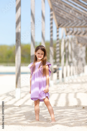 Obraz na płótnie Carefree summer. Little girl in lilac dress has fun and posing on sandy beach under wooden gazebo on sunny day. Joyful childhood adventures and tropical vacation concept. Space for text. w salonie