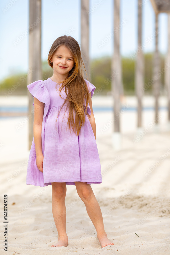 Carefree summer. Little girl in lilac dress has fun and posing on sandy beach under wooden gazebo on sunny day. Joyful childhood adventures and tropical vacation concept. Space for text.