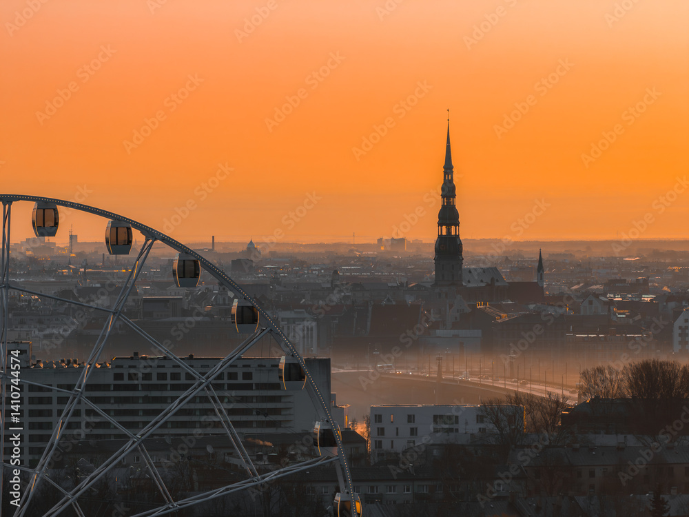 Serene sunrise over Riga, Latvia, featuring St. Peter's Church spire, a Ferris wheel in the foreground, and a warm orange sky with soft morning light.