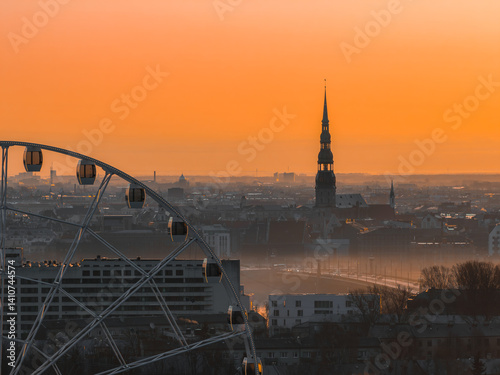 Obraz na płótnie Serene sunrise over Riga, Latvia, featuring St. Peter's Church spire, a Ferris wheel in the foreground, and a warm orange sky with soft morning light. w salonie