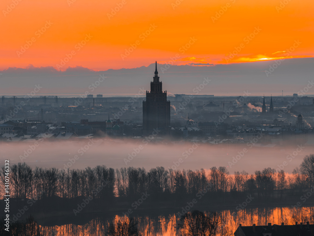 A vibrant sunrise over Riga, Latvia, with the Latvian Academy of Sciences in view, mist covered cityscape, and the Daugava River reflecting warm hues.