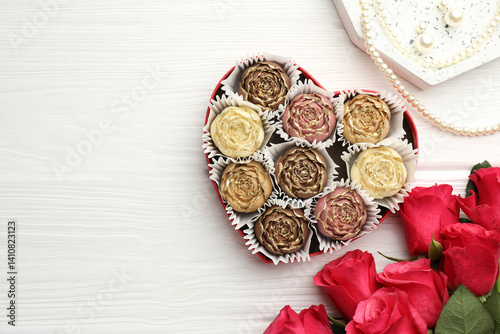 Flower shaped chocolate bonbons in box, red roses and pearl jewelry on white wooden table, flat lay. Space for text