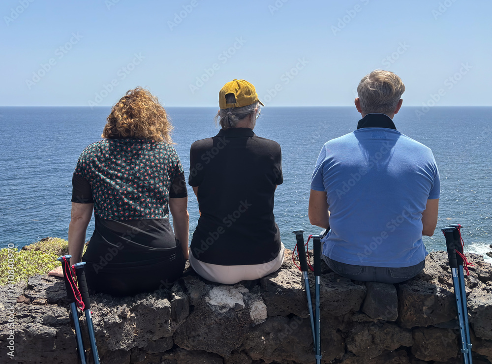 Fototapeta premium Rear view of three bonding friends sitting face the sea in a trekking sunny day admiring the horizon over water