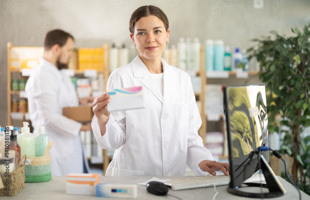 Fototapeta premium Young woman pharmacist with package of pills, she stands behind the counter near the computer. Pharmacist works in a pharmacy against the background of an male assistant who puts goods on the shelves