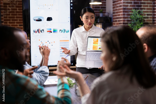 Female entrepreneur discusses financial planning with employees, using laptop to display business analytics. The evening meeting highlights readiness, strategy and deadline driven decision making. photo