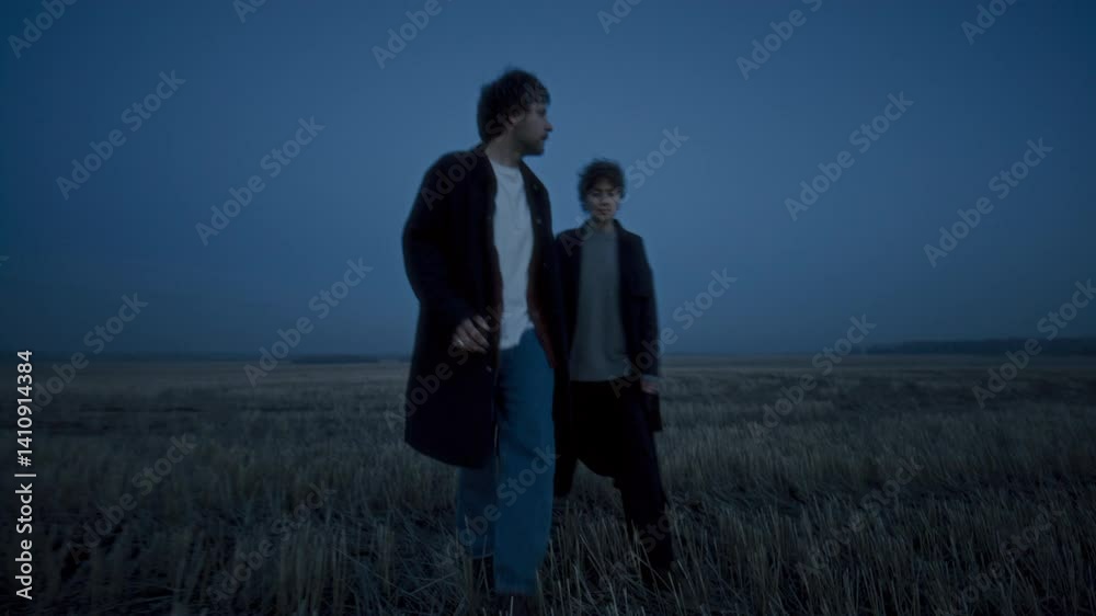 Young couple walking hand in hand across dry grassy field at dusk under blue evening sky, looking around at rural landscape Young couple walking hand in hand across dry grassy field at dusk under blue evening sky, looking around at rural landscape