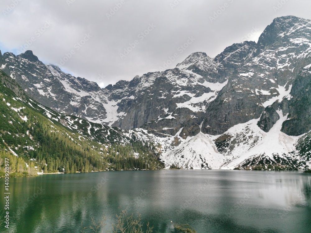 Naklejka premium Snow-covered Tatra Mountains surround the emerald waters of Morskie Oko lake, with dense pine forests and dramatic rocky slopes under cloudy skies