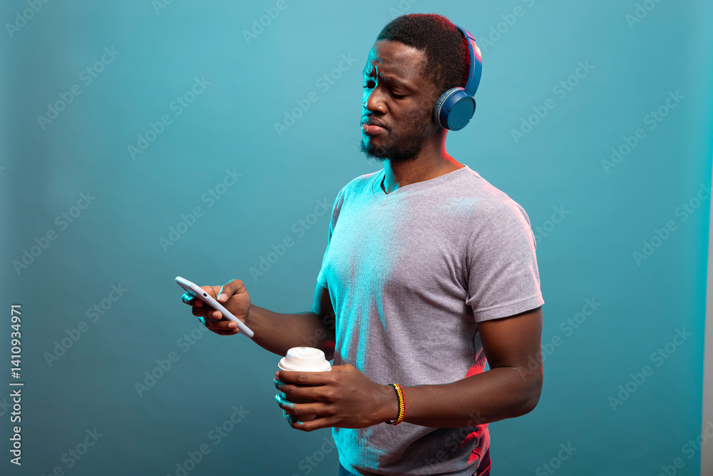 Young male individual holding a disposable cup and looking at his cellphone with a confused expression. Black man with wireless headphones unsure with a video on his mobile device.