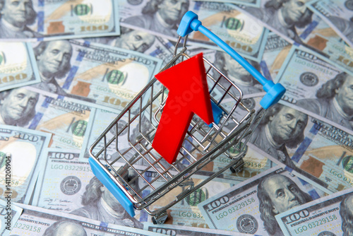 A shopping cart and red upward pointing arrow on a bed of blurred one hundred U.S. dollar bills. Increasing costs, inflation, or rising consumer spending concept.
