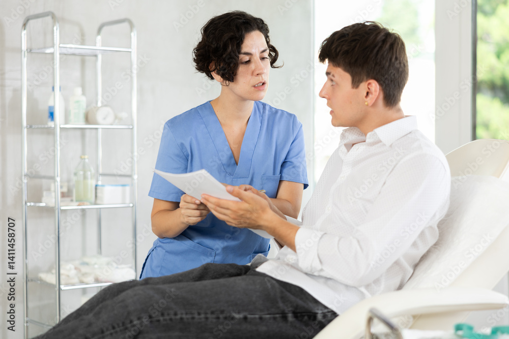 Young male patient lying at examination couch reading and signing documents while talking with female doctor Young male patient lying at examination couch reading and signing documents while talking with female doctor