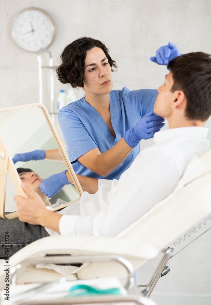 Young man sits in chair at a doctor appointment with beautician. Woman doing facial examination before surgery