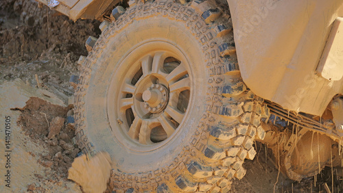 A close-up of a heavily mud-covered off-road vehicle tire with deep tread. The tire is partially submerged in mud, showcasing its rugged design and ability to handle challenging terrain.
