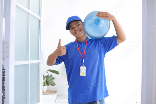 Foto Smiling Young Asian Delivery Man In Blue Uniform Carrying Gallon And Giving Thum