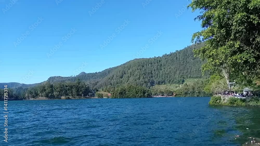 Video from the edge of Sarangan Lake in Indonesia on a bright morning. Blue sky, green mountain range, calm water waves. A lake in the mountainous area of Indonesia. Video from the edge of Sarangan Lake in Indonesia on a bright morning. Blue sky, green mountain range, calm water waves. A lake in the mountainous area of Indonesia.