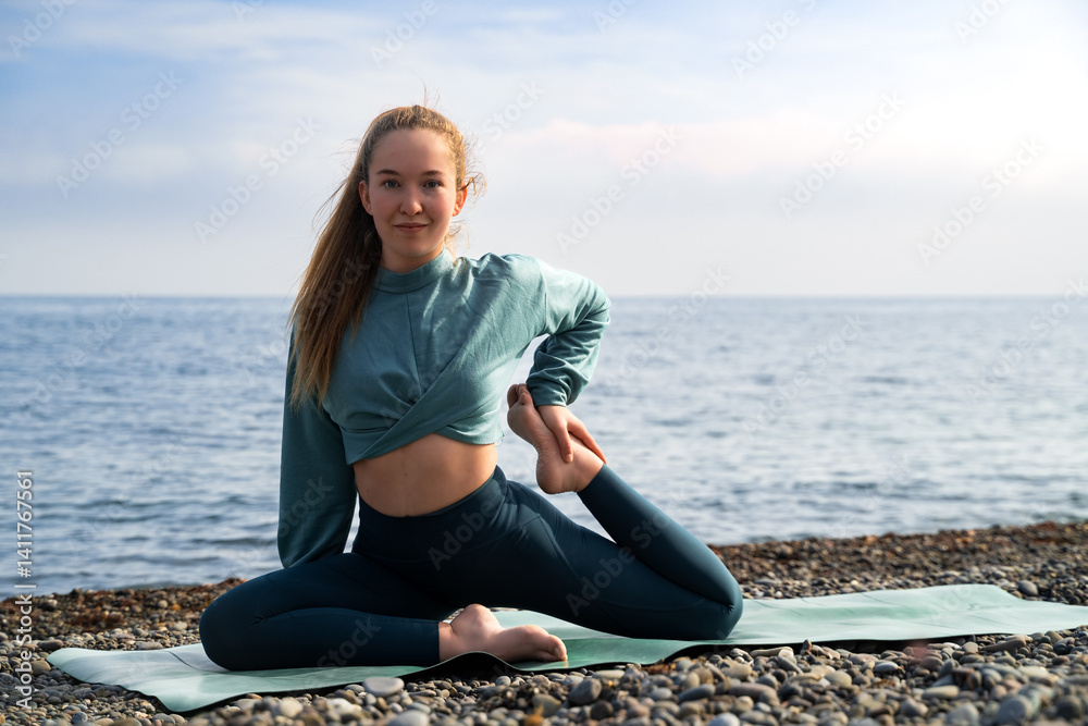 Young woman practicing yoga exercises on a fitness mat, doing mermaid pose, wearing activewear, looking at the camera, smiling, feeling wellness, stretching, working out on a sunny day on the seashore Young woman practicing yoga exercises on a fitness mat, doing mermaid pose, wearing activewear, looking at the camera, smiling, feeling wellness, stretching, working out on a sunny day on the seashore