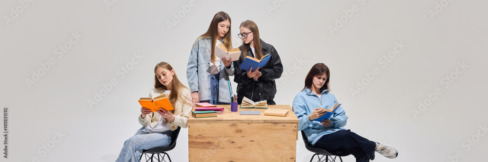 Group of teenage girls in classroom, wearing casual outfits and actively participating in reading tasks, doing homework together against grey background. Concept of modern schooling, education