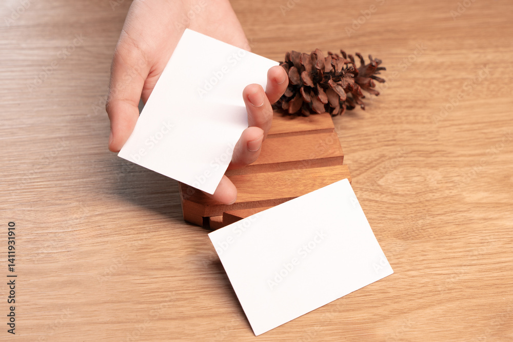 Obraz premium Hand holding white blank business card mockup with wooden decoration on brown background