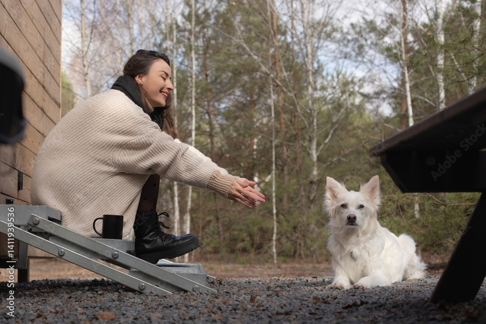Woman in cozy beige sweater and black boots is sitting on the steps of wooden cabin, reaching out to her white dog, who is calmly lying nearby.Serene outdoor forest background adds peaceful atmosphere