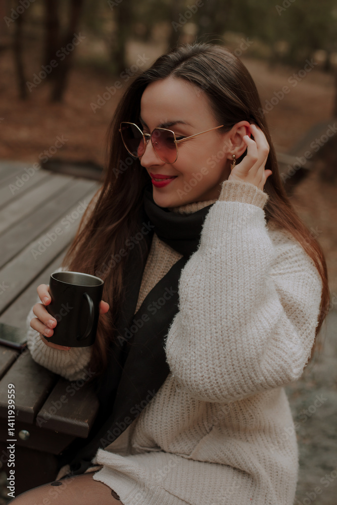 A woman sits at a wooden table outdoors, smiling while holding a black mug. She wears sunglasses, a beige sweater, and a black scarf. The background shows a peaceful forest setting.
