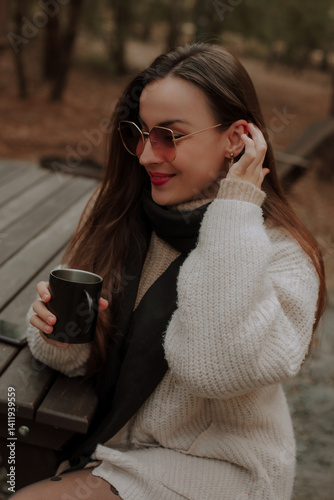 Obraz na płótnie A woman sits at a wooden table outdoors, smiling while holding a black mug. She wears sunglasses, a beige sweater, and a black scarf. The background shows a peaceful forest setting. w salonie